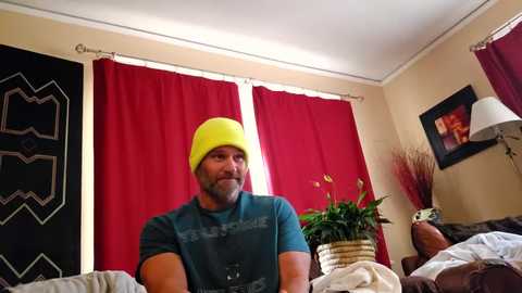 Video of a bearded man in a yellow beanie, blue t-shirt, and glasses, sitting on a couch in a cozy room with red curtains, potted plant, and framed photo on beige wall.