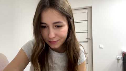 A video of a young Caucasian woman with long, straight brown hair and fair skin, wearing a grey T-shirt, smiling softly. The background shows a white door and part of a bathroom, with a hint of a towel rack.