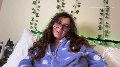Video of a smiling, fair-skinned woman with curly brown hair, wearing glasses, a blue polka dot robe, and sitting in a cozy room with fairy lights and a white pillow.