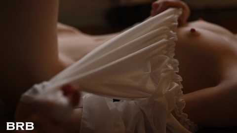 Video: Close-up of a woman with fair skin, topless, holding up a white, ruffled dress with her hands. Background is dimly lit, focusing on the fabric.