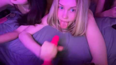 Video of a blonde woman with a blue tongue licking a red vibrator, surrounded by two other women on a bed, bathed in purple light.