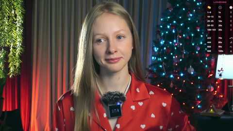 Video of a blonde woman in a red shirt with white hearts, microphone, smiling, Christmas tree in background.