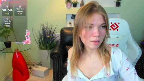 A video of a young woman with light skin and straight blonde hair, wearing a white off-shoulder shirt, sitting in a gaming chair in a green-walled room.