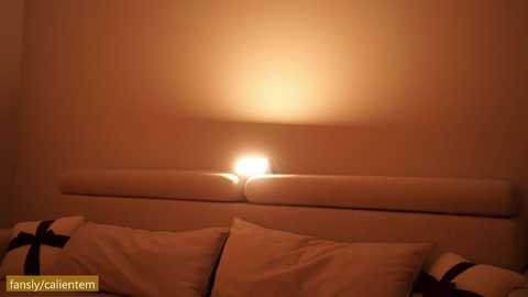 Media: A video of a dimly lit bedroom featuring a beige wall, a white headboard with a lit lamp, and a bed with white pillows and a blue and white striped pillow. The scene is warmly lit, creating a cozy atmosphere.