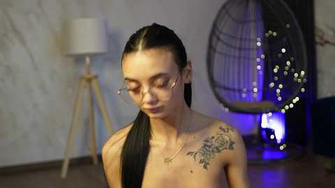 Video of a topless woman with long, dark hair, wearing glasses, and intricate floral tattoo on her shoulder. She stands in a dimly lit room with a hanging chair and modern lamp.