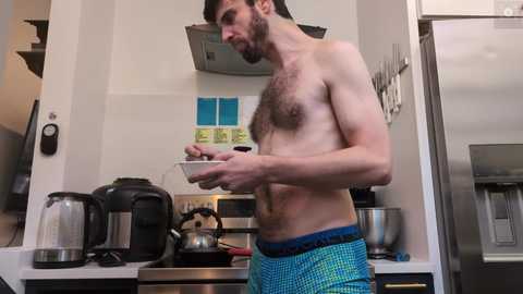 Video of a shirtless, muscular man with a beard in blue plaid shorts, holding a plate in a modern kitchen with stainless steel appliances, white cabinets, and blue-tiled backsplash.