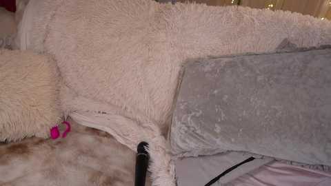Video of a plush, beige-fur throw with a pink heart-shaped toy and a grey, plush pillow, both on a beige bedspread.