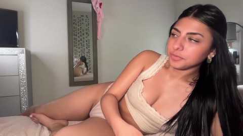 Video of a young Latina woman with long black hair, wearing a beige bra and panties, posing seductively on a bed in a softly lit room.