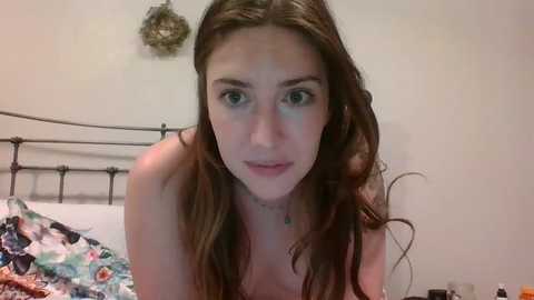 A video of a young Caucasian woman with long brown hair, light skin, and blue eyes, leaning forward, wearing a floral-patterned top, in a bedroom with a metal bed frame and a floral blanket.