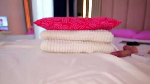 Video of a neatly stacked pile of pillows on a bed, featuring a pink and white striped pillow on top, set against a softly lit, modern bedroom with white walls and a TV visible in the background.