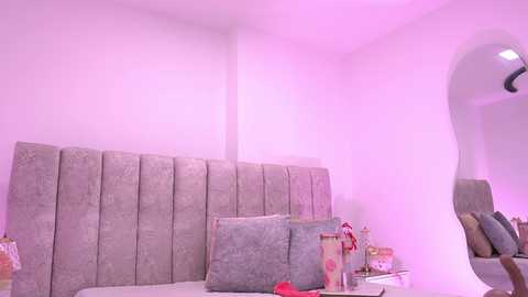 A video of a modern, minimalist bedroom with a plush, gray upholstered headboard, soft pastel pink lighting, and scattered decorative pillows in various shades. The room's clean, white walls and a sleek, curved mirror add to its contemporary ambiance.