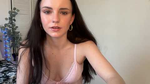 Video of a fair-skinned woman with long dark hair, wearing a light pink lace bralette, sitting in a room with white walls, a closed window, and a black-and-white patterned blanket.