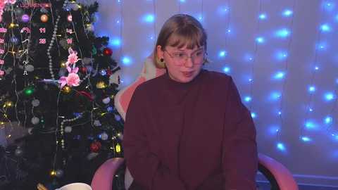 Video of a young girl with glasses and short brown hair, wearing a maroon sweater, sitting in a pink chair in front of a lit Christmas tree adorned with blue and white fairy lights.
