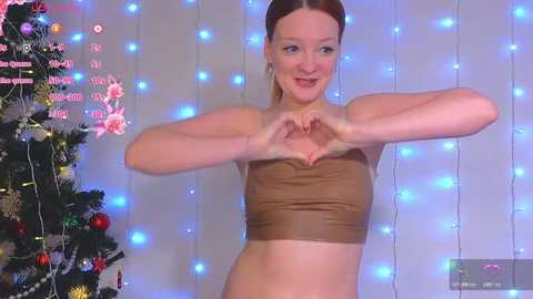 Video of a smiling, fair-skinned woman with red hair, making a heart shape with her hands in front of a Christmas tree adorned with blue fairy lights and ornaments.