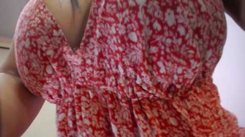 A close-up video of a woman's upper torso in a red floral-patterned dress, showcasing her large breasts and defined waist. The background is out of focus, featuring a light-colored wall.