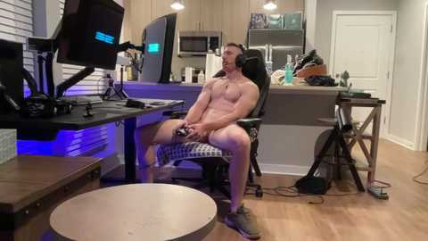 Video of a muscular, naked man with a gaming headset, seated in a gaming chair, playing a video game in a modern, well-lit kitchen with wooden floors and sleek cabinetry.