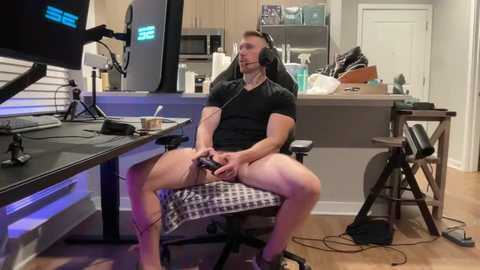 Video of a muscular man with short hair, wearing a black t-shirt, seated on a gaming chair in a modern kitchen, holding a controller, surrounded by gaming equipment.