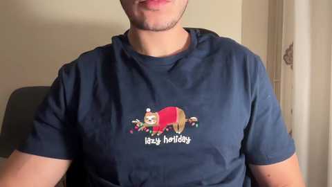 Video of a man wearing a navy blue t-shirt with a festive Christmas-themed graphic featuring a red Santa Claus and a snowman, \"Merry Holidays\" printed in white text. The background shows a beige wall and a partially visible door.