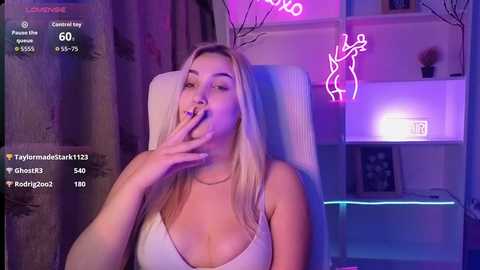 Video of a blonde woman with long hair, wearing a white tank top, biting her finger, in a neon-lit room with pink and purple hues.