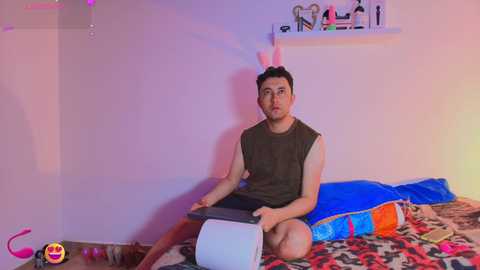 Video of a young man with a pink bunny headband, sitting on a bed with a leopard-patterned blanket, wearing a sleeveless shirt and shorts, surrounded by colorful pillows.