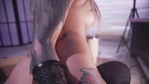 Video of a woman with light skin, long dark hair, and tattoos, partially undressed, in a dimly lit room with purple lighting, sitting on a chair.