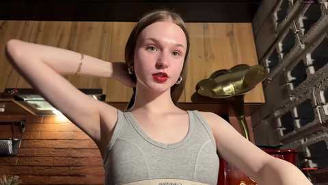Media: Video of a fair-skinned, red-lipped woman with straight, light brown hair, wearing a gray sports bra and standing in a rustic kitchen with wooden walls and metal shelving.