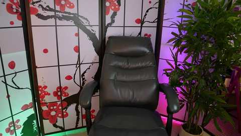 A video of a black leather recliner chair with armrests, set against a backdrop of a folding screen with red cherry blossom designs and a green potted plant on the right.