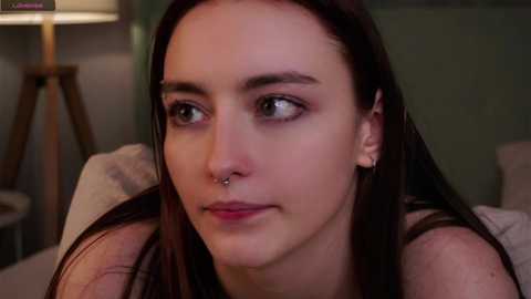 Video of a young woman with long dark hair, fair skin, and a septum piercing, wearing a black top, gazing thoughtfully. Background includes a lamp and beige furniture.