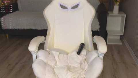 Video of a white gaming chair with a beige blanket draped over it, a black controller placed on the seat, and a small white stand beside it.