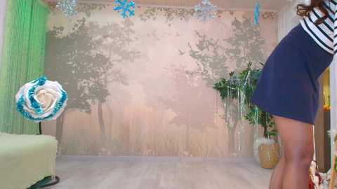 Video of a young woman in a dark blue skirt and white top facing a painted mural of a forest. Background includes a green chair, a potted plant, and decorative ornaments.