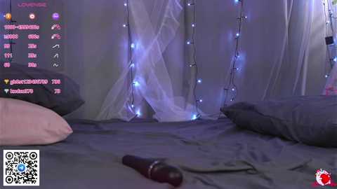 Video of a dimly lit bedroom with a bed covered in dark gray sheets, fairy lights, and a pink pillow. A black vibrator rests on the bed.