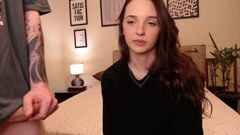 Video of a young woman with long brown hair, wearing a black shirt, sitting on a bed in a cozy, beige-themed room with a lamp, framed art, and potted plants.
