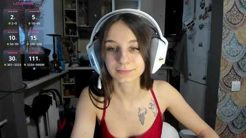 Video of a young woman with light skin and straight brown hair, wearing a red spaghetti strap top and large white headphones, sitting in a cluttered room with a virtual overlay showing streaming stats.