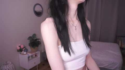 Media: Video of a slender, fair-skinned woman with long, black hair, wearing a white crop top, standing in a softly lit bedroom. Background includes a bed, white nightstand with potted plants, and a wall-mounted circular mirror.