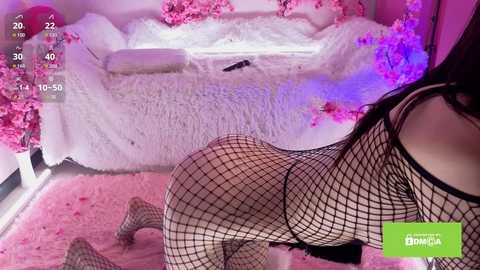 A video of a woman with long dark hair, wearing a black fishnet bodysuit, posing provocatively on a pink shag carpet in a room decorated with white fur and pink cherry blossom petals.