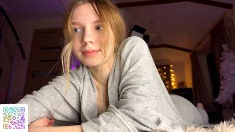 Video of a young Caucasian woman with blonde hair, lying on her stomach in a cozy bedroom, wearing a grey robe. The room has warm lighting and a soft, inviting atmosphere.