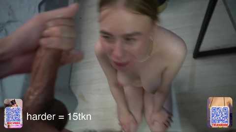 Video of a naked, light-skinned woman with small breasts, kneeling on a tiled floor, looking up at a large, erect penis held by a hand.