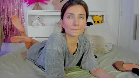 Video of a young woman with light skin and brown hair, lying on a bed in a playful pose, wearing a gray t-shirt, surrounded by stuffed toys and a pink tinsel backdrop.