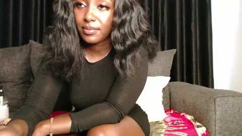 Video of a Black woman with dark skin, long wavy black hair, and a slender build, wearing a black sheer top, seated on a gray couch with red and white pillows, against black curtains.