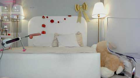 Video of a modern, minimalist bedroom with a white bed, yellow bow, red heart decorations, plush teddy bear, and two white lamps.