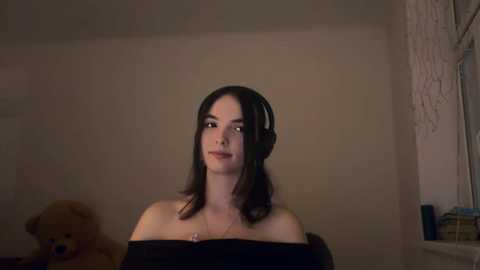 A young woman with fair skin, straight dark hair, and a slim build, wearing black headphones, stands in a dimly lit room with beige walls and a teddy bear.