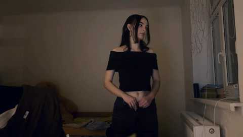 Video of a slender, dark-haired woman in a black off-shoulder crop top and black pants, standing in a dimly lit, messy bedroom with a window and a cluttered dresser.
