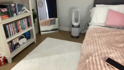 Media: Video of a modern, minimalist bedroom with a plush beige rug, white bed with pink pillow, gray wall, white shelves with books, and a white air purifier.