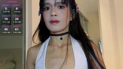 Video of an Asian woman with long black hair, wearing a white halter top, black choker, and black hair clips, indoors with a digital screen showing weather and temperature details.