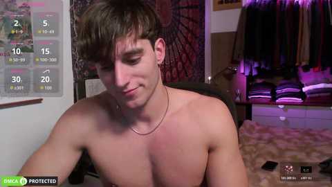 Video of a shirtless, pale-skinned, young man with short brown hair, wearing a thin silver chain necklace, sitting on a chair, looking down. Background shows a cluttered room with a bed, a closet, and a wall calendar.