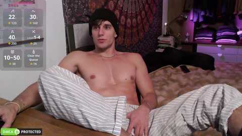 Video of a shirtless, light-skinned, muscular young man with short dark hair, wearing striped pajama pants, reclining on a bed in a dimly lit room with a TV and a patterned tapestry in the background.