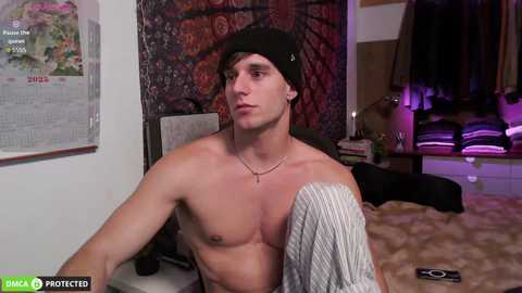 Video of a shirtless young man with a black beanie, sitting on a bed, wearing a necklace, in a dimly lit room with a colorful tapestry and a calendar on the wall.