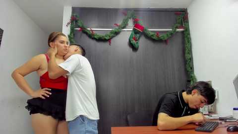 Video of a man passionately kissing a woman in a red top, black shorts, while a man in a black shirt works on a computer in a Christmas-decorated office.