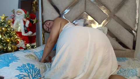 Video of a fair-skinned woman with short hair, wearing a sheer white dress, crawling on a bed with a festive blue and white blanket. Background includes a large, ornate headboard, a Christmas tree with gold ornaments, and a Santa Claus figurine.