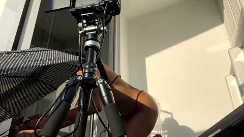 Video of a professional videographer with a tripod-mounted camera, capturing a woman in a black bikini on a sunlit balcony, with a modern, minimalistic background.